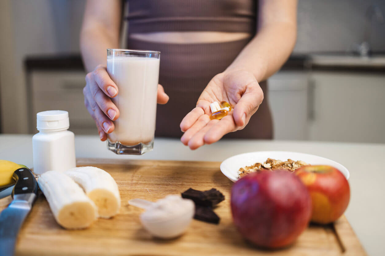 Pessoa segurando cápsulas de suplementos para colesterol ao lado de um copo de leite, frutas e cereais sobre a mesa, representando cuidados com a saúde